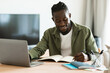 © Prostock-studio - Smiling african american man sitting at desk, working on laptop and taking notes in notebook, black male studying online