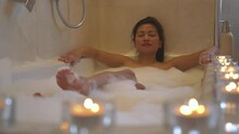 Attractive Lady With Eyes Closed Chilling In A Bubbly Bath Embraced With Candles. Young Asian Woman Relaxing In Bathtub. Lined Up Glowing Candles For Relaxing Atmosphere And Pleasant Spa Experience.