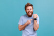 © khosrork - Portrait of positive funny bearded man standing with open mouth and satisfied expression, pointing with finger ar smartwatch and laughing. Indoor studio shot isolated on blue background.