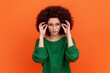 Portrait of attentive beautiful woman with Afro hairstyle wearing green casual style sweater standing holding glasses and looking at camera. Indoor studio shot isolated on orange background.