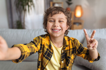 Selfie time. Camera pov portrait of happy little boy blogger making photo, smiling to camera and showing peace gesture