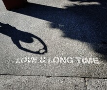 Grafitti Street Art On The Footpath Of The Words Love You Long Time With A Girl's Shadow Signing A Heart With Her Hands.
