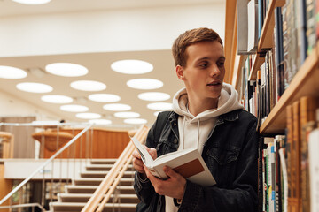 Wall Mural - Young student with book looking at bookshelf. Teenager in library reading a book.