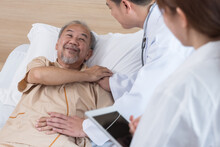 Asian Doctor And Nurse Visit And Take Care Senior Man Patient In Room. Attractive Two Therapist Practitioner Explain Diagnosis In Hospital Ward To Treatment To Elderly Older Male During Appointment.