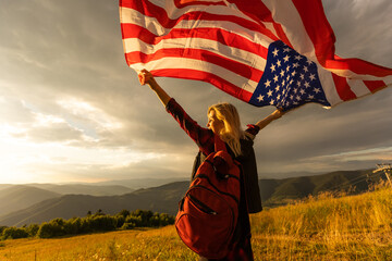 Young happy american woman with long hair holding waving on wind USA national flag on her sholders relaxing outdoors enjoying warm summer day