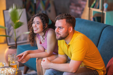 At home in living room attractive couple watching a favourite sport match after their team getting the victory they are hugging each other and are excited lady eating some snacks