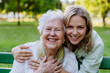 © Halfpoint - Portrait of adult granddaughter hugging her senior grandmother when sitting on bench in park.
