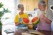 Happy senior caucasian couple in home kitchen holding a big and heavy red watermelon cutted in a half - husband with thumb up - hydration, freshness, diet and healthy eating concept