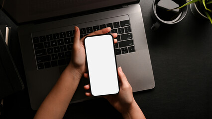 A female hands holding a smartphone white screen mockup over modern black office desk
