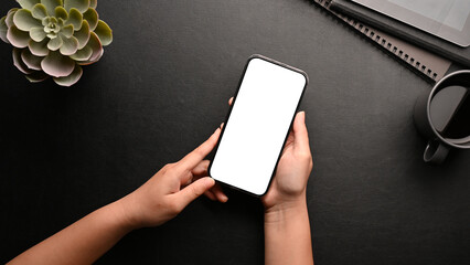Top view, A female hands holding a smartphone white screen mockup over black office desk.