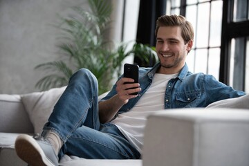 attractive smiling young man wearing casual clothes sitting on a couch at the living room, using mob