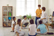 © Studio Romantic - Children and teacher having circle time in class. Group of happy, cheerful school students sitting on the floor in a modern classroom interior, listening to interesting stories and discussing them