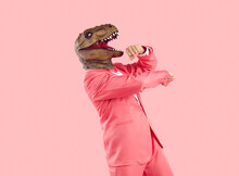 Cheerful Funny And Humorous Man In Dinosaur Rubber Mask Dances Isolated On Pastel Pink Background. Young Man In Formal Pink Wears Absurd Animal Mask On His Head. Creative Concept For Advertising,