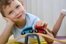 Close-up Of A Smiling Boy With Blond Hair Lying Down Playing With Wooden Toys On Puzzle Mats. A Child Of 4-5 Years Old Thinks How To Ride A Red Train, A Blue Locomotive Or Wagons On A Wooden Railway.