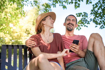 Cheerful young couple smiling and laughing using smart phone sitting in a bench in a park. Man and woman friends sharing media using a phone together outside with love.