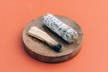 Burning incense bundle. White sage and burning palo santo stick on wooden stand over orange brown background. Fragrant bundle for meditation and room fumigation. Selective focus