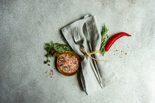 Overhead View Of A Rustic Cutlery Setting On A Napkin With Pink Himalayan Salt With Fresh Rosemary, Thyme And Chilli Pepper