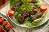 Fototapeta Kuchnia - Traditional south european skinless sausages cevapcici made of ground meat and spices on lettuce leaves with thyme and tomatoes on white plate on kitchen towel, black silverware