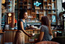 Happy Black Waitress Serving Coffee To Female Guest In Cafe.