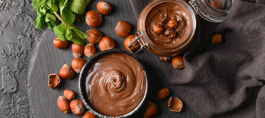 Bowl and jar with tasty chocolate paste and hazelnuts on dark background, top view