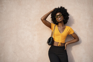 Wall Mural - Happy African young woman with afro hair wearing stylish sunglasses posing against beige wall 