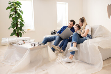 Young family of three sitting on sofa in room undergoing renovation. Middle-aged brunette father, dressed in blue jeans white t-shirt and checkered shirt shows wife and child design plan for new home.