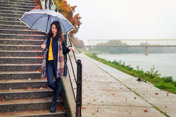 Wall Mural - Young woman with umbrella walking in the rain beside river