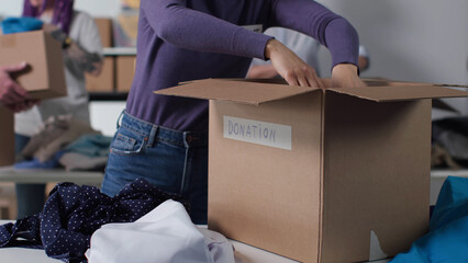 Wall Mural - Close up of female hands filling cardboard donation box with clothes at warehouse. 