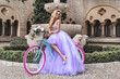 Elegant luxury fashion. Glamour, stylish elegant woman in amazing long gown dress on colorful bike. Female model in lilac long dress in the city. Travel model. Outdoor shoot. Vogue. Couture.