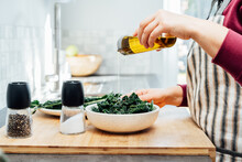 Close Up Woman Adding Olive Oil While Cooking Kale Chips Or Healthy Salad For Dinner On The Kitchen Table. Healthy Eating, Dieting Lifestyle. Selective Focus, Copy Space.