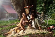 Picnic Of Couple Man And Woman Sitting On Rug Under Tree In Park Over Vintage Bicycle Background. Reading Book. Romantic Concept. Have Free Time Out Together During Weekend In Summer.
