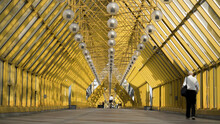 People Walking Inside Pedestrian Bridge In Moscow, Russia. Action. View Inside Of Andreyevskiy Bridge With Bright Yellow Metal Beams And Hanging Lanterns.