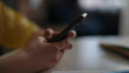 Close-up hands of unrecognizable child boy scrolling touching screen with fingers on smartphone sitting at table. Kid fingers scrolling and tapping touch screen cellphone using mobile app.