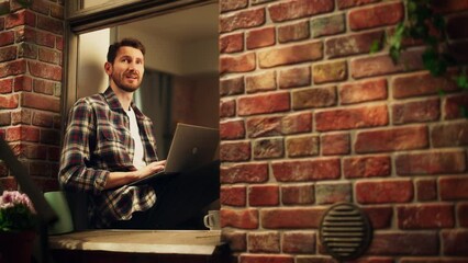 Aufkleber - Handsome Creative Male Works on a Laptop Computer while Sitting on a Windowsill of his Cozy Apartment. Professional Working Remotely from Home, Listens Birds Sing. Static Shot Through Building Window.