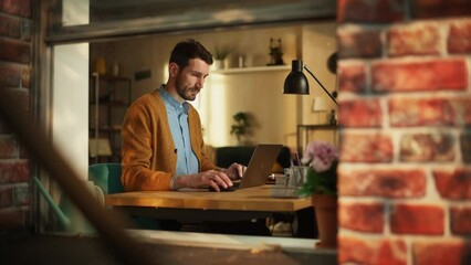 Leinwandbilder - Male Specialist Working on Laptop Computer in Stylish Apartment. Creative Man Managing Corporate Social Media, Browsing Internet. Freelancer Remote Work From Home. View From Outdoors into Window.