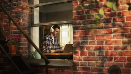 Leinwandbilder - Modern Freelancer Typing on a Laptop, Listens Music in Headphones. Concept of Professional e-Business Entrepreneur at Home Office. View From Outdoors into Window.