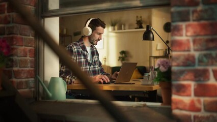 Aufkleber - Handsome Caucasian Man Working on Laptop Computer while Sitting Behind His Desk at Home Office. Freelance Entrepreneur Listening to Music, Doing Remote Telework. View From Outdoors into Window.