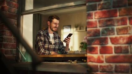 Leinwandbilder - Happy Smiling Man Sitting Behind Desk at Home Office and Using Smartphone. Handsome Man Browsing Through Internet, Checking Videos on Social Media, Shopping Online. View From Outdoors into Window.