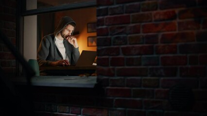 Aufkleber - Information Technology Specialist Working Late on a Latptop. Software Engineer Wearing Hoodie, Working on Computer in His Cozy Home Office Apartment. Remote Work. Night View From Outdoors into Window.