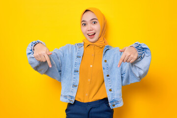 Surprised young Asian woman in jeans jacket pointing fingers down, inviting customers to special event isolated over yellow background