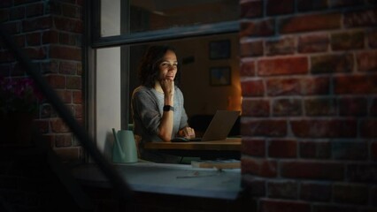 Leinwandbilder - Smiling Hispanic Woman at Home Using Laptop on the Warm Cozy Evening. Professional Freelancer Working Remotely From Home. Shot From Outside Into the Window of Apartment.