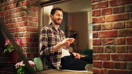 Leinwandbilder - Happy Handsome Caucasian Man Using Smartphone while Sitting on a Windowsill of his Cozy Apartment. Casual Shirt Wearing Guy Browsing Internet, Checking Videos on Social Media, Having Fun on Sunny Day.