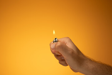 Lit cigarette lighter and flame held in hand by Caucasian male hand. Close up studio shot, isolated on orange background