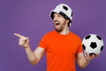Young Fun Fan Man He In Orange T-shirt Cheer Up Support Football Sport Team Hold Soccer Ball Panama Hat Watch Tv Live Stream Point Index Finger Aside On Workspace Isolated On Plain Purple Background.