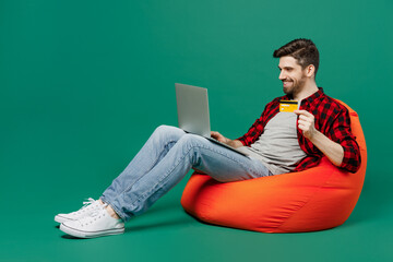 Poster - Full body young man he in red shirt grey t-shirt sit in bag chair using laptop pc computer hold credit bank card do online shopping order delivery booking tour isolated on plain dark green background.