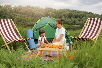 Wall Mural - Outdoor shot of smiling satisfied young adult woman and little girl sitting on blanket near tent, having picnic near river, enjoying their resting together.