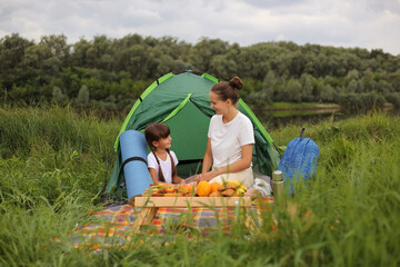 Wall Mural - Image of pleased pretty mother having picnic near river with her daughter, sitting on plaid on the ground near tent, talking and enjoying their camping.