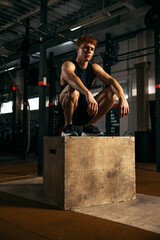 Wall Mural - Portrait of sportive, muscular red-haired young man sitting on box, resting isolated over gym background