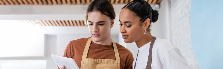 Young interracial sellers in aprons using digital tablet in confectionery, banner.