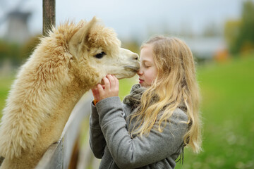 Cute young girl stroking an alpaca at a farm zoo on autumn day. Child feeding a llama on an animal farm. Kid at a petting zoo at fall.